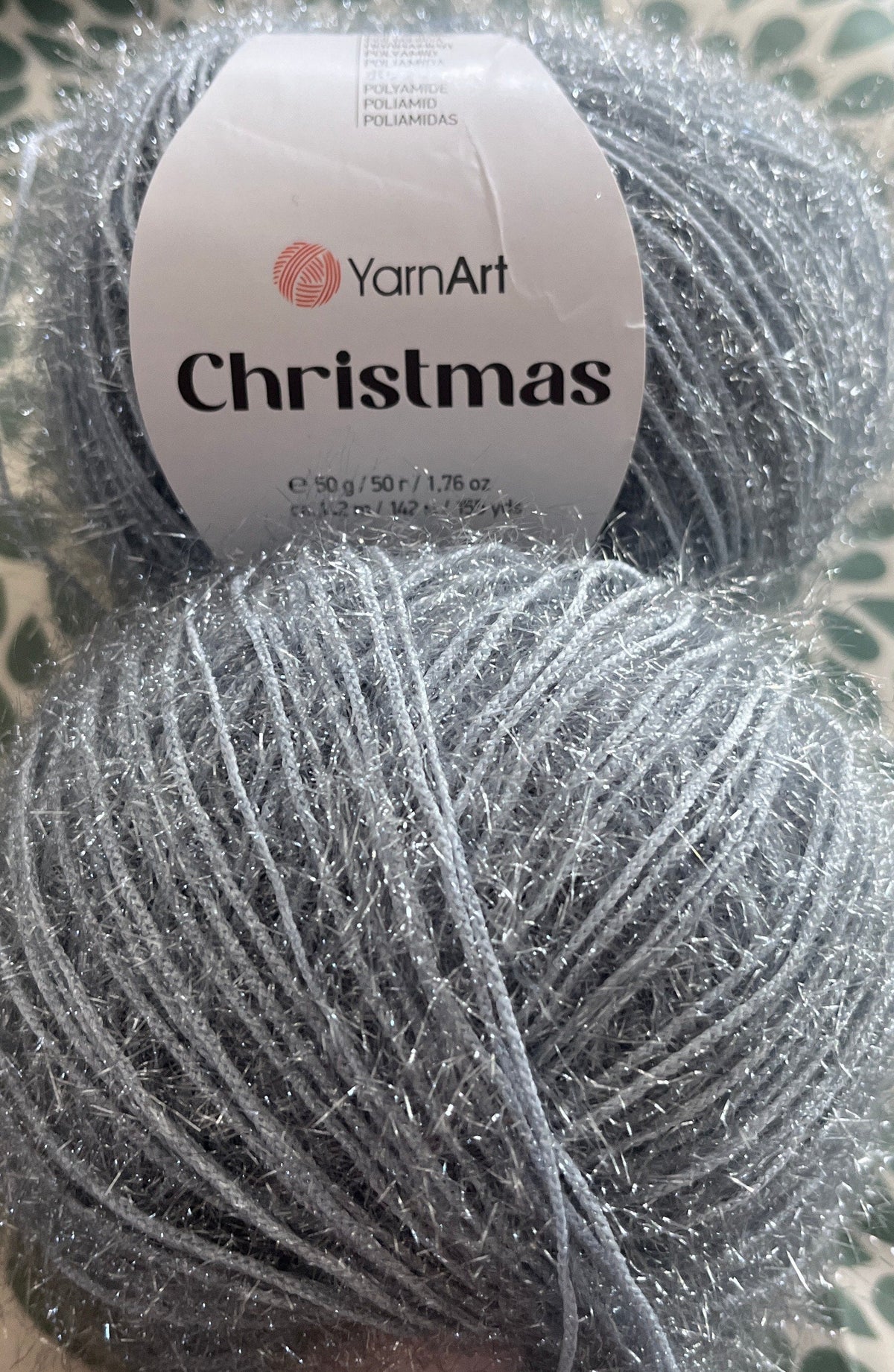A ball of sparkly silver-gray yarn with a white label reading 'YarnArt Christmas' resting on a green and white patterned fabric surface.